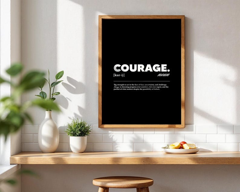 May include: A framed black and white print with the word "COURAGE" in bold white letters. Below the word is the pronunciation and the definition of courage. The frame is a warm brown color, and the print is displayed on a white wall.
