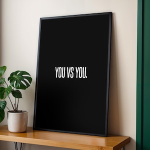 Peut inclure: Un cadre noir avec le texte blanc "YOU VS YOU.". Il est posé sur une surface en bois, à côté d'une plante en pot avec de grandes feuilles vertes. Le fond est un mur neutre et une porte verte.