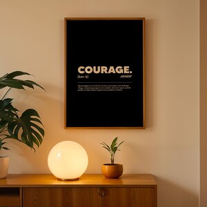 May include: A framed black poster with the word "COURAGE" in white, along with a definition. The poster is in a wooden frame and hangs on a beige wall. A round, glowing lamp and a small potted plant sit on a wooden cabinet below.