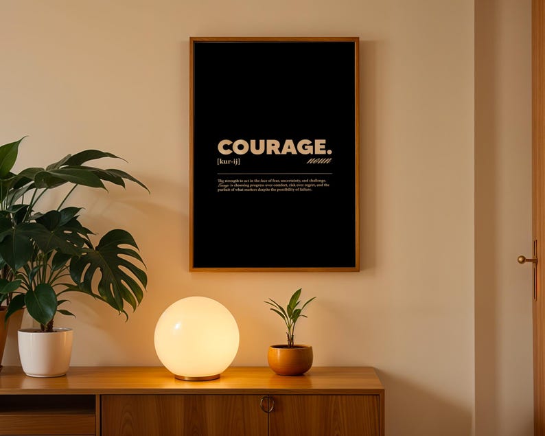 May include: A framed black poster with the word "COURAGE" in white, along with a definition. The poster is in a wooden frame and hangs on a beige wall. A round, glowing lamp and a small potted plant sit on a wooden cabinet below.