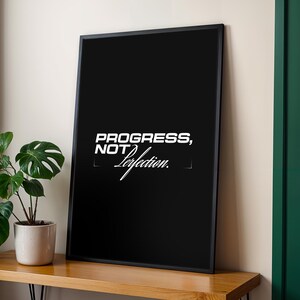 Może przedstawiać: Czarny, oprawiony plakat z białym napisem "PROGRESS, NOT Perfection". Plakat jest wyświetlany na drewnianej powierzchni. Obok plakatu znajduje się roślina doniczkowa z zielonymi liśćmi. Rama jest czarna, a tło ma neutralny kolor.
