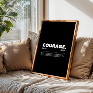 May include: A framed black print with the word "COURAGE" in white, along with a definition. The wooden frame is a light brown color. The print is propped on a beige sofa with matching pillows, near a window with a plant.