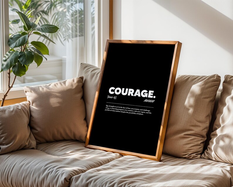May include: A framed black print with the word "COURAGE" in white, along with a definition. The wooden frame is a light brown color. The print is propped on a beige sofa with matching pillows, near a window with a plant.