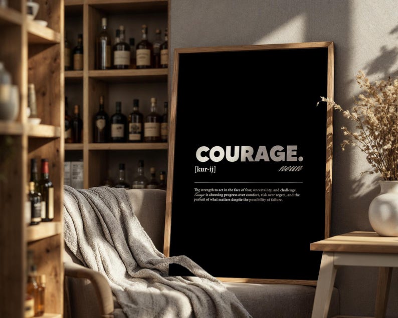 May include: A framed black print with the word "COURAGE" in white, with the definition below. The print is displayed in a room with a wooden bookshelf and a neutral-colored blanket draped over a chair.