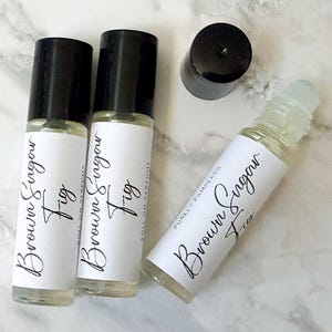 May include: Three roll-on perfume bottles with black lids and white labels. The labels read "Brown Sugar Fig" in elegant script. One lid is off, and the bottles are arranged on a marble-patterned surface. The bottles are labeled "Purely Pampered".