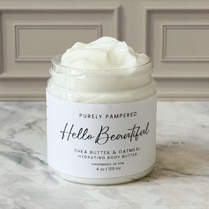 Puede incluir: Un tarro de vidrio transparente lleno de manteca corporal blanca. El tarro tiene una etiqueta blanca con el texto "PURELY PAMPERED Hello Beautiful SHEA BUTTER & OATMEAL HYDRATING BODY BUTTER HANDMADE IN USA 4 oz | 120 ml". La manteca corporal es de color blanco cremoso.