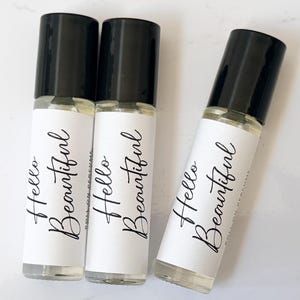 Puede incluir: Tres frascos de perfume roll-on de vidrio con tapas negras. Cada frasco tiene una etiqueta blanca con las palabras "Hello Beautiful" en elegante escritura negra. Los frascos están llenos de un líquido transparente.