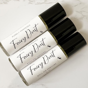 May include: Three roll-on perfume bottles with black caps and white labels. The labels read "Fairy Dust" in cursive script, with the words "Purely Pampered" and "Roll on Perfume" also printed. Each bottle contains 10 ml of perfume.