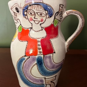 May include: A white ceramic pitcher with a whimsical, hand-painted design. The artwork features a figure in a red jacket, blue hat, and purple pants. The handle and rim have a glittery gold trim. A unique piece for serving beverages.