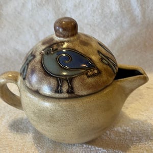 May include: A ceramic creamer with a lid. The creamer is beige with a brown handle and spout. The lid is brown with a blue and green bird design. The bird has a blue body and green wings. The creamer is round and has a small handle.