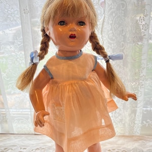May include: A vintage doll with blonde braided pigtails tied with blue ribbons. The doll wears a peach-colored dress with a blue trim and matching shoes. The doll is standing on a black stand.