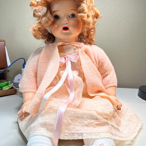 May include: A vintage doll with curly red hair, blue eyes, and a surprised expression. The doll wears a peach-colored dress, sweater, and white socks with floral accents. Pink and white ribbons adorn the dress.