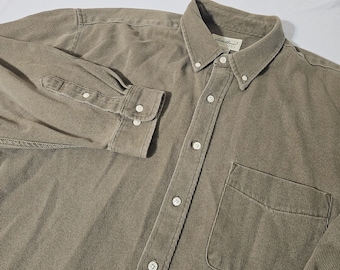 Puede incluir: Una camisa abotonada de manga larga en un color verde oliva apagado. La camisa presenta una textura de pana, un cuello abotonado, un bolsillo en el pecho y puños abotonados. La etiqueta Eddie Bauer es visible.