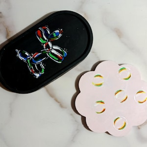 Puede incluir: Bandeja ovalada negra con un diseño de perro globo colorido. También hay un posavasos rosa claro en forma de flor con diseños circulares coloridos. Ambos artículos están sobre una superficie de mármol.