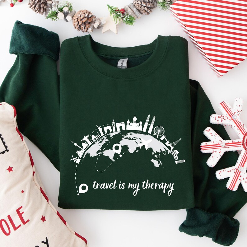 May include: A dark green sweatshirt with a white graphic of a world map and famous landmarks. The text "travel is my therapy" is printed below the map. The sweatshirt has dark green cuffs and a crew neck.