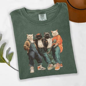 Puede incluir: Camiseta verde Comfort Colors con un gr&aacute;fico de cuatro gatos vestidos con ropa urbana. Los gatos llevan chaquetas, petos y zapatillas deportivas. La camiseta tiene el logotipo de Comfort Colors. Prenda de vestir informal.