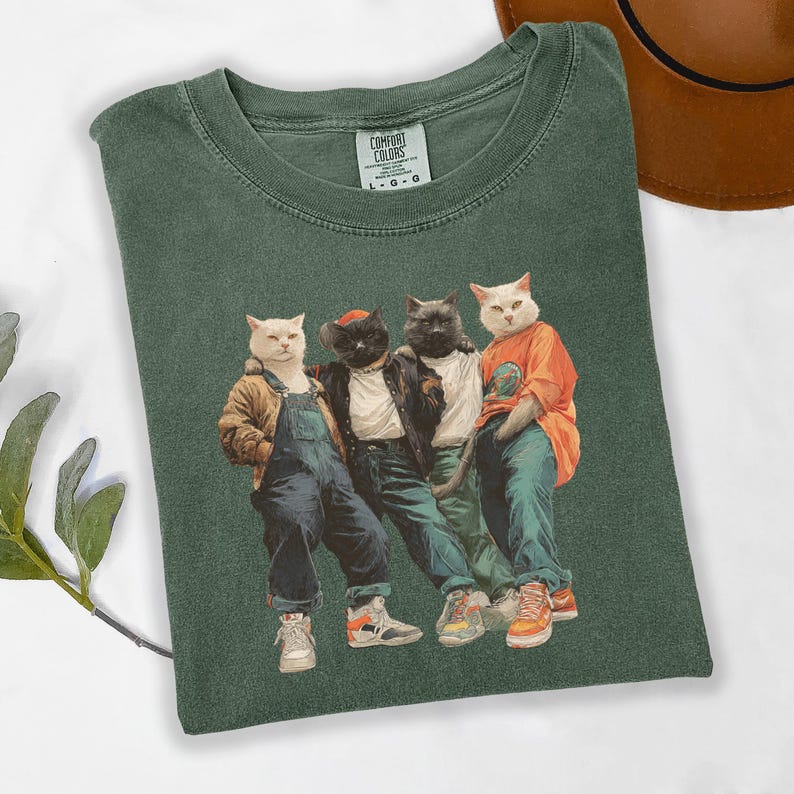 Puede incluir: Camiseta verde Comfort Colors con un gr&aacute;fico de cuatro gatos vestidos con ropa urbana. Los gatos llevan chaquetas, petos y zapatillas deportivas. La camiseta tiene el logotipo de Comfort Colors. Prenda de vestir informal.