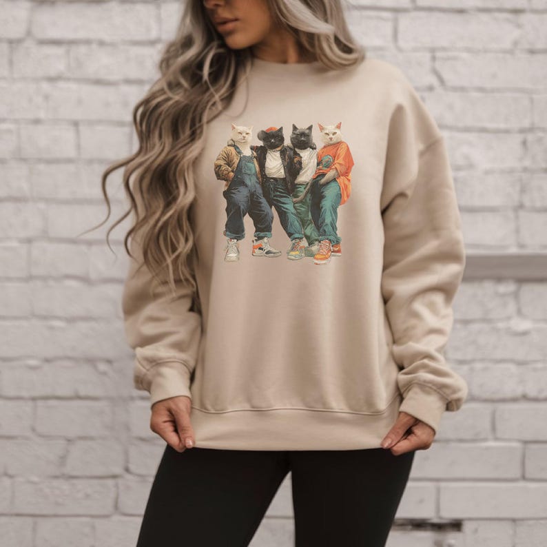 Puede incluir: Sudadera beige de cuello redondo con un gr&aacute;fico de cuatro gatos vestidos con ropa urbana. Los gatos llevan chaquetas, petos y zapatillas deportivas. La sudadera es de color neutro, adecuada para uso casual.
