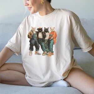 Puede incluir: Camiseta color crema con un gr&aacute;fico de cuatro gatos vestidos con ropa urbana. Los gatos llevan chaquetas, petos y zapatillas deportivas. La imagen tiene una est&eacute;tica vintage, ligeramente descolorida, con los gatos en varias poses.