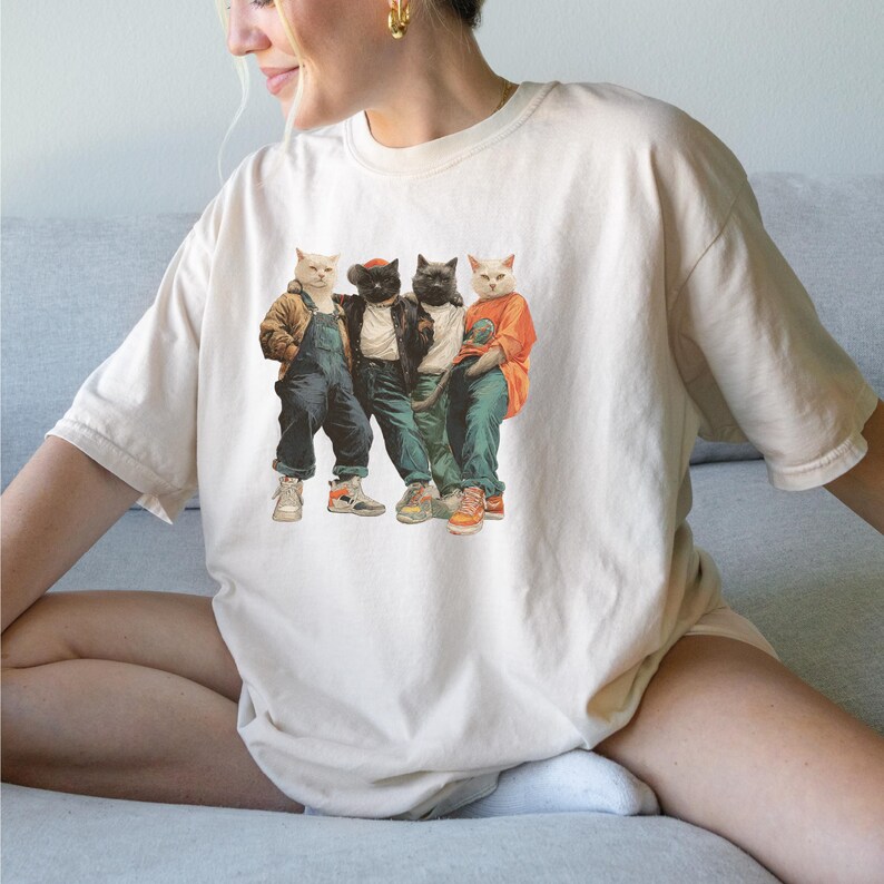 Puede incluir: Camiseta color crema con un gr&aacute;fico de cuatro gatos vestidos con ropa urbana. Los gatos llevan chaquetas, petos y zapatillas deportivas. La imagen tiene una est&eacute;tica vintage, ligeramente descolorida, con los gatos en varias poses.