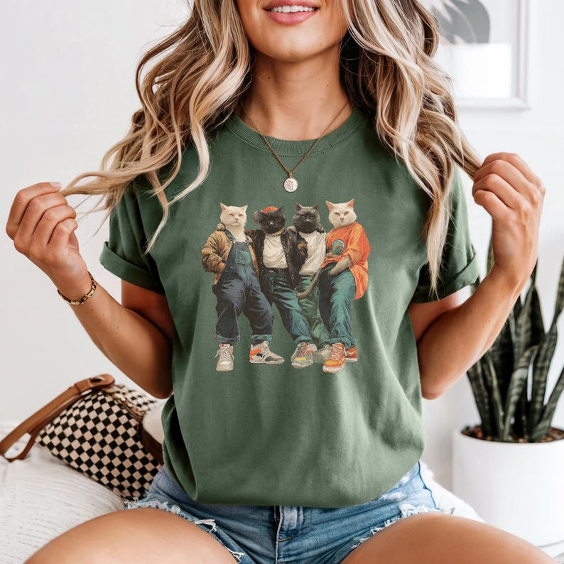 Puede incluir: Camiseta verde con un gr&aacute;fico de cuatro gatos vestidos con ropa urbana. Los gatos llevan chaquetas, petos y zapatillas deportivas. El dise&ntilde;o tiene una est&eacute;tica vintage y retro.