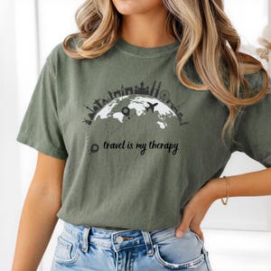 May include: Olive green t-shirt featuring a black and white graphic of a world map with city skylines and a plane. The text "travel is my therapy" is printed below the graphic. The shirt is made of a soft material.