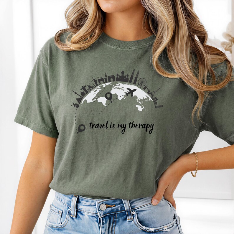 May include: Olive green t-shirt featuring a black and white graphic of a world map with city skylines and a plane. The text "travel is my therapy" is printed below the graphic. The shirt is made of a soft material.