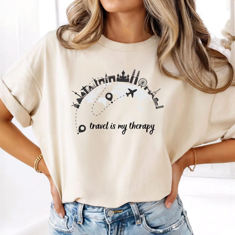May include: Beige t-shirt with a black graphic of world landmarks and a dotted line with a plane. The text "travel is my therapy" is below the graphic. The shirt is made of a soft material.