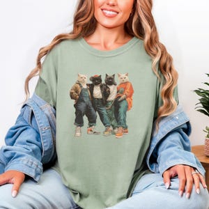 Puede incluir: Camiseta verde salvia con un gr&aacute;fico de cuatro gatos vestidos con ropa urbana. Los gatos llevan monos, una chaqueta de cuero y una camisa naranja. Los gatos est&aacute;n juntos, con una est&eacute;tica vintage.