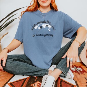 May include: A blue t-shirt with a graphic of a world map, airplane, and city skylines. The text "travel is my therapy" is printed below the graphic. The shirt is a solid color and has short sleeves.