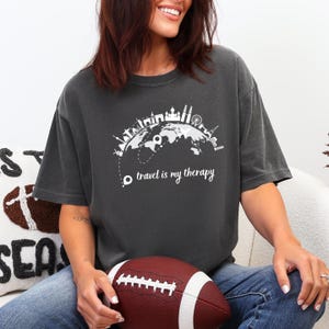 May include: Dark grey t-shirt with a white graphic of a globe featuring famous landmarks and the text "travel is my therapy." The shirt is a relaxed fit and has short sleeves. A brown football is held in the lap.