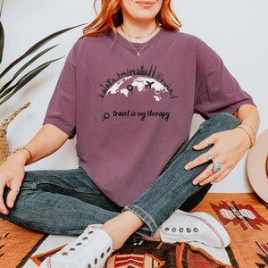 May include: A maroon t-shirt with a graphic of a world map, airplane, and city skylines. The text "travel is my therapy" is printed below the graphic. The shirt is a casual style.