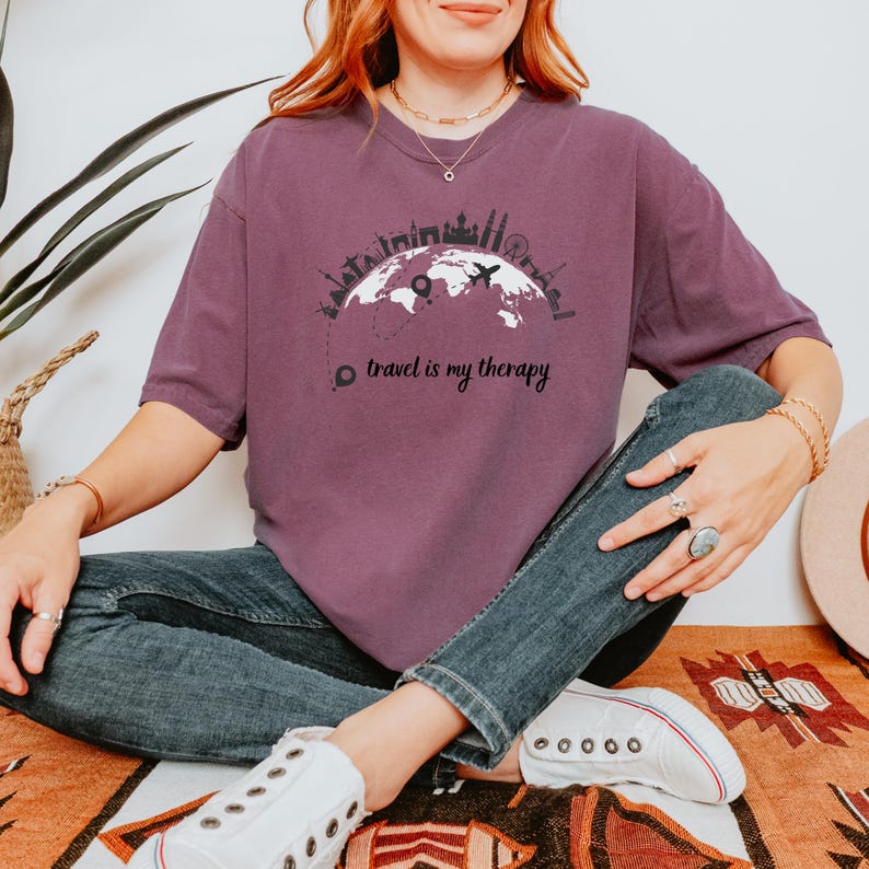 May include: A maroon t-shirt with a graphic of a world map, airplane, and city skylines. The text "travel is my therapy" is printed below the graphic. The shirt is a casual style.