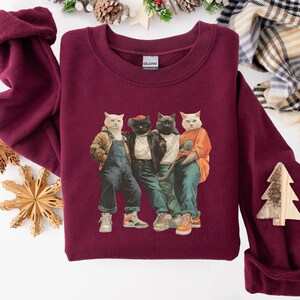 Puede incluir: Sudadera burdeos de cuello redondo con un gr&aacute;fico de cuatro gatos vestidos con ropa informal. Los gatos llevan una chaqueta, un peto, una chaqueta de cuero y una camiseta naranja. La sudadera tiene un corte cl&aacute;sico y est&aacute; hecha de un material suave.