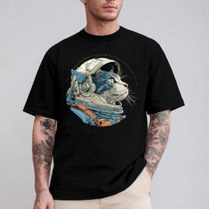May include: Black t-shirt featuring a detailed illustration of a cat wearing an astronaut helmet. The cat is depicted in shades of blue, gray, and white, with orange straps. The design is set against a black background.