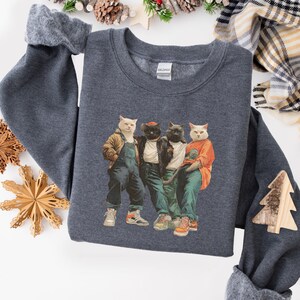 Puede incluir: Una sudadera gris oscuro con un gr&aacute;fico de cuatro gatos vestidos con ropa humana. Los gatos llevan chaquetas, petos y zapatillas deportivas. La sudadera tiene cuello redondo y mangas largas.