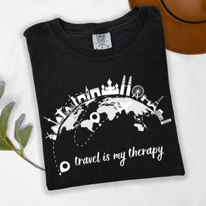 May include: Black t-shirt with a white graphic of a globe featuring famous landmarks and the text "travel is my therapy." The shirt is made of a soft material and is a comfortable fit. A brown hat is visible in the background.