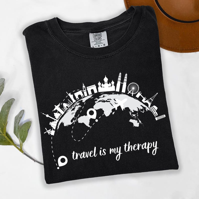 May include: Black t-shirt with a white graphic of a globe featuring famous landmarks and the text "travel is my therapy." The shirt is made of a soft material and is a comfortable fit. A brown hat is visible in the background.