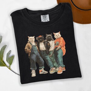 Puede incluir: Camiseta negra con un gr&aacute;fico de cuatro gatos vestidos con ropa urbana. Los gatos llevan chaquetas, monos y zapatillas deportivas. La camiseta tiene el logo de Comfort Colors en la esquina superior izquierda.