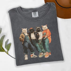 Puede incluir: Camiseta Comfort Colors gris oscuro con un gr&aacute;fico de cuatro gatos vestidos con ropa urbana. Los gatos llevan monos, una chaqueta de cuero y una camiseta naranja. La camiseta tiene cuello redondo y mangas cortas.