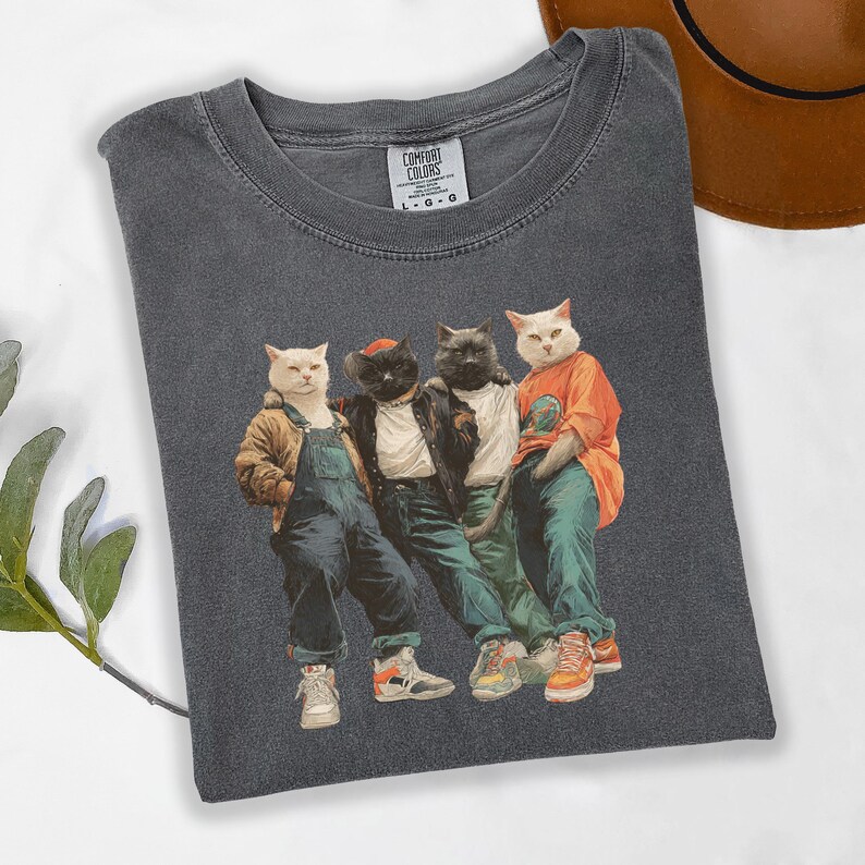 Puede incluir: Camiseta Comfort Colors gris oscuro con un gr&aacute;fico de cuatro gatos vestidos con ropa urbana. Los gatos llevan monos, una chaqueta de cuero y una camiseta naranja. La camiseta tiene cuello redondo y mangas cortas.