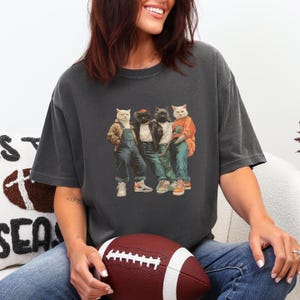Puede incluir: Camiseta gris oscuro con un dise&ntilde;o de cuatro gatos vestidos con ropa urbana. Los gatos llevan chaquetas, petos y zapatillas deportivas. La imagen tiene un aspecto vintage, ligeramente descolorido.