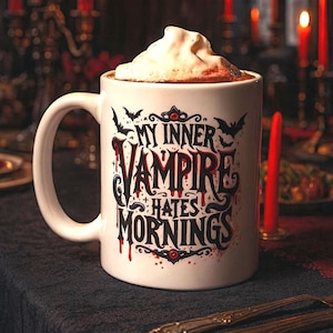 Puede incluir: Taza de cer&aacute;mica blanca con el texto "MY INNER VAMPIRE HATES MORNINGS" en fuente g&oacute;tica, llena de una bebida oscura y cubierta con crema batida. La taza est&aacute; sobre una mesa con velas rojas encendidas y un mantel oscuro.