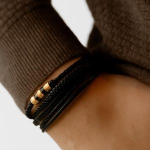 May include: A stack of black leather bracelets with gold-colored metal accents. The bracelets are worn on a person's wrist, and the image is taken from a close-up angle. The person is wearing a brown sweater.