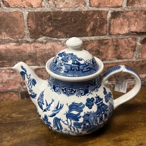 May include: A white ceramic teapot featuring a blue willow pattern with detailed illustrations of pagodas, willow trees, and birds. The teapot includes a matching lid with a knob handle, a curved spout, and a handle with a small white tag attached.
