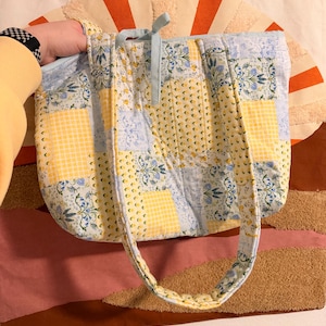 May include: A quilted tote bag featuring a patchwork design of light blue and yellow floral patterns mixed with yellow and white gingham squares. The bag includes two padded shoulder straps and a light blue fabric tie closure at the top center.