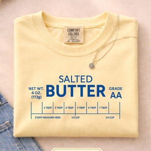May include: A light yellow t-shirt with the words "SALTED BUTTER" in blue. The shirt includes a measurement guide for butter, with a net weight of 4 oz (113g). A silver necklace with a flower pendant rests on the shirt.