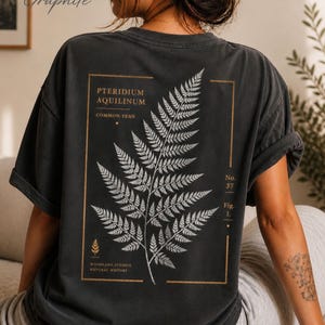 May include: A graphite-colored t-shirt featuring a botanical print on the back. The graphic displays a white fern illustration within a gold rectangular frame, accompanied by the text Pteridium Aquilinum, Common Fern, No. 37, Fig. 1, and Woodland Studies Natural History.
