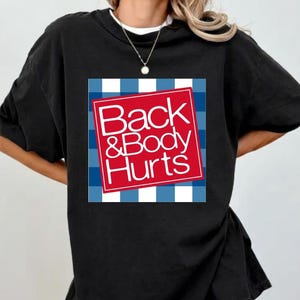 May include: Black t-shirt with a graphic that reads "Back & Body Hurts" in white text on a red square, set against a blue and white checkered background. The shirt has short sleeves and a crew neck.