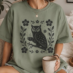 Könnte beinhalten: Salbeigrünes T-Shirt mit einem schwarz-weißen Eulen-Design, das eine Kaffeetasse hält, umgeben von floralen und Blättermotiven in einem quadratischen Rahmen. Das T-Shirt hat eine lockere Passform.